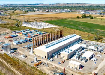 Aerial view of an industrial complex with transformers, equipment yards, and storage containers near open fields and distant town hillsides.
