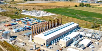 Aerial view of an industrial complex with transformers, equipment yards, and storage containers near open fields and distant town hillsides.