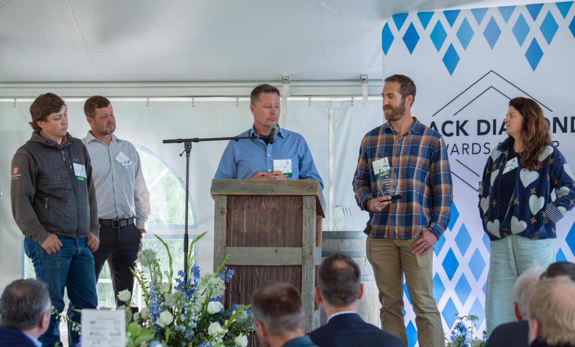 Man at a wooden lectern speaks into a microphone while four others stand beside him on a stage at an event with name badges visible on their shirts/lamps.