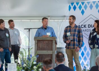 Man at a wooden lectern speaks into a microphone while four others stand beside him on a stage at an event with name badges visible on their shirts/lamps.