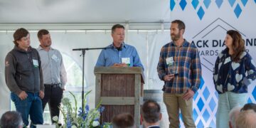 Man at a wooden lectern speaks into a microphone while four others stand beside him on a stage at an event with name badges visible on their shirts/lamps.