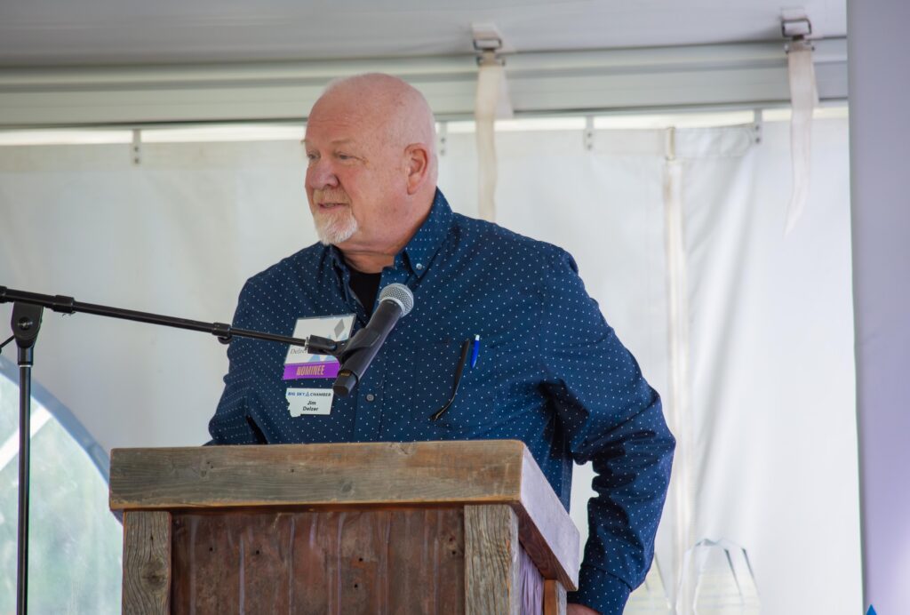 Older man with a white beard speaking into a microphone at a wooden podium, wearing a dark blue polka-dot shirt and event badge (nominee).