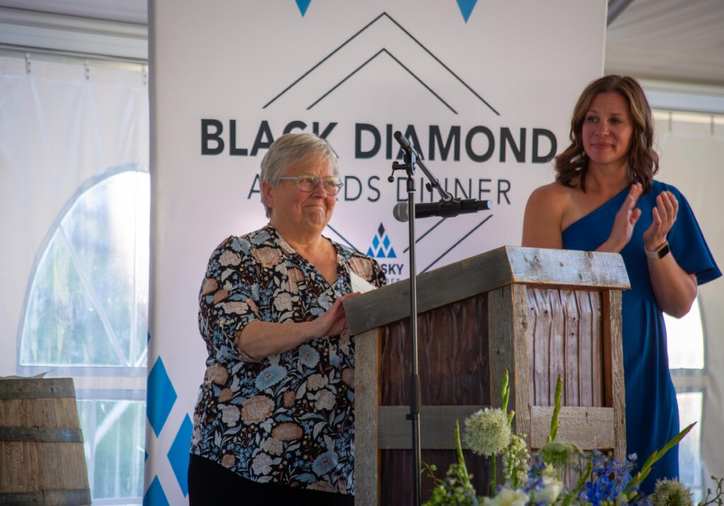 Older woman in a floral blouse speaking at a wooden podium, microphone in front, with a banner behind reading 'BLACK DIAMOND AWARDS DINNER' and a woman in blue beside her.
