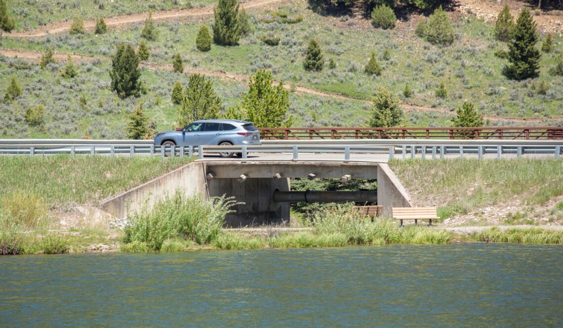 Silver SUV crossing a bridge over a river, with grassy, tree-filled hills in the background