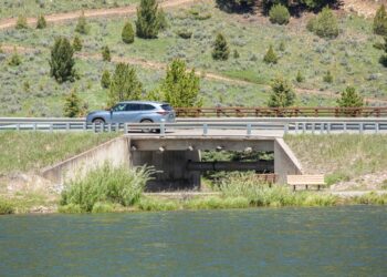 Silver SUV crossing a bridge over a river, with grassy, tree-filled hills in the background