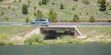 Silver SUV crossing a bridge over a river, with grassy, tree-filled hills in the background