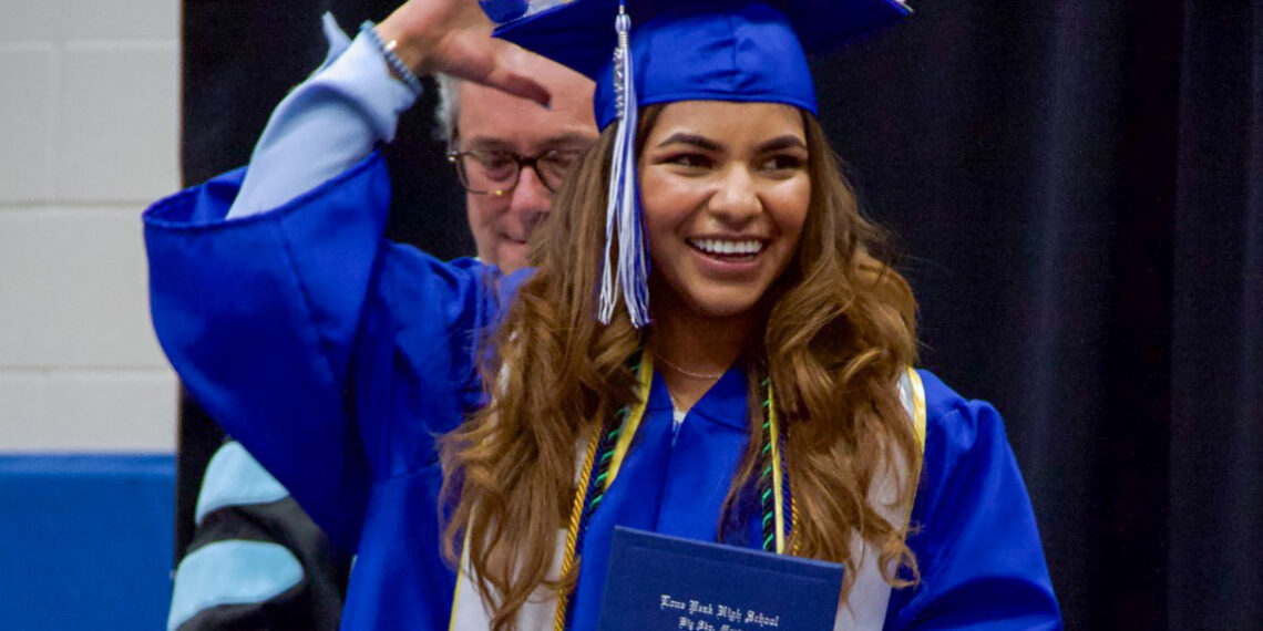 Smiling graduate in a blue cap and gown holds a diploma during a commencement ceremony.