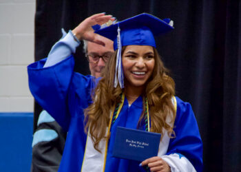Smiling graduate in a blue cap and gown holds a diploma during a commencement ceremony.