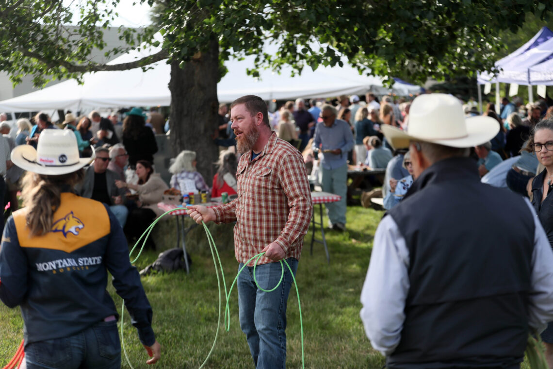 Man in a plaid shirt holding a green rope lasso in the foreground at a crowded outdoor festival with white tents and trees behind.