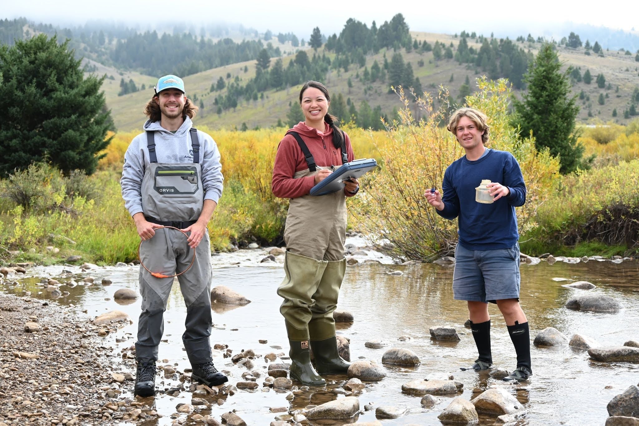 Three hikers stand in a shallow rocky river, smiling, with forested hills in the background.