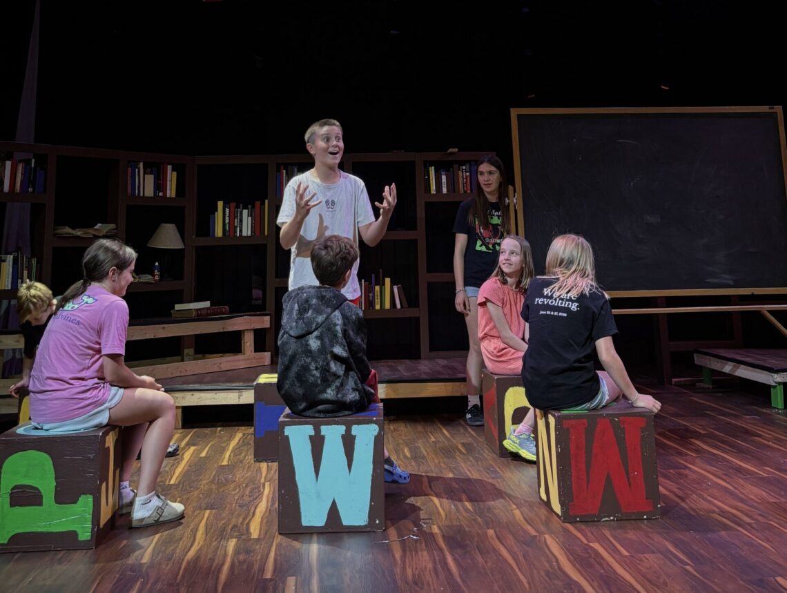 Group of kids on a classroom-style stage with oversized colorful alphabet blocks and bookshelves in the background under stage lights.