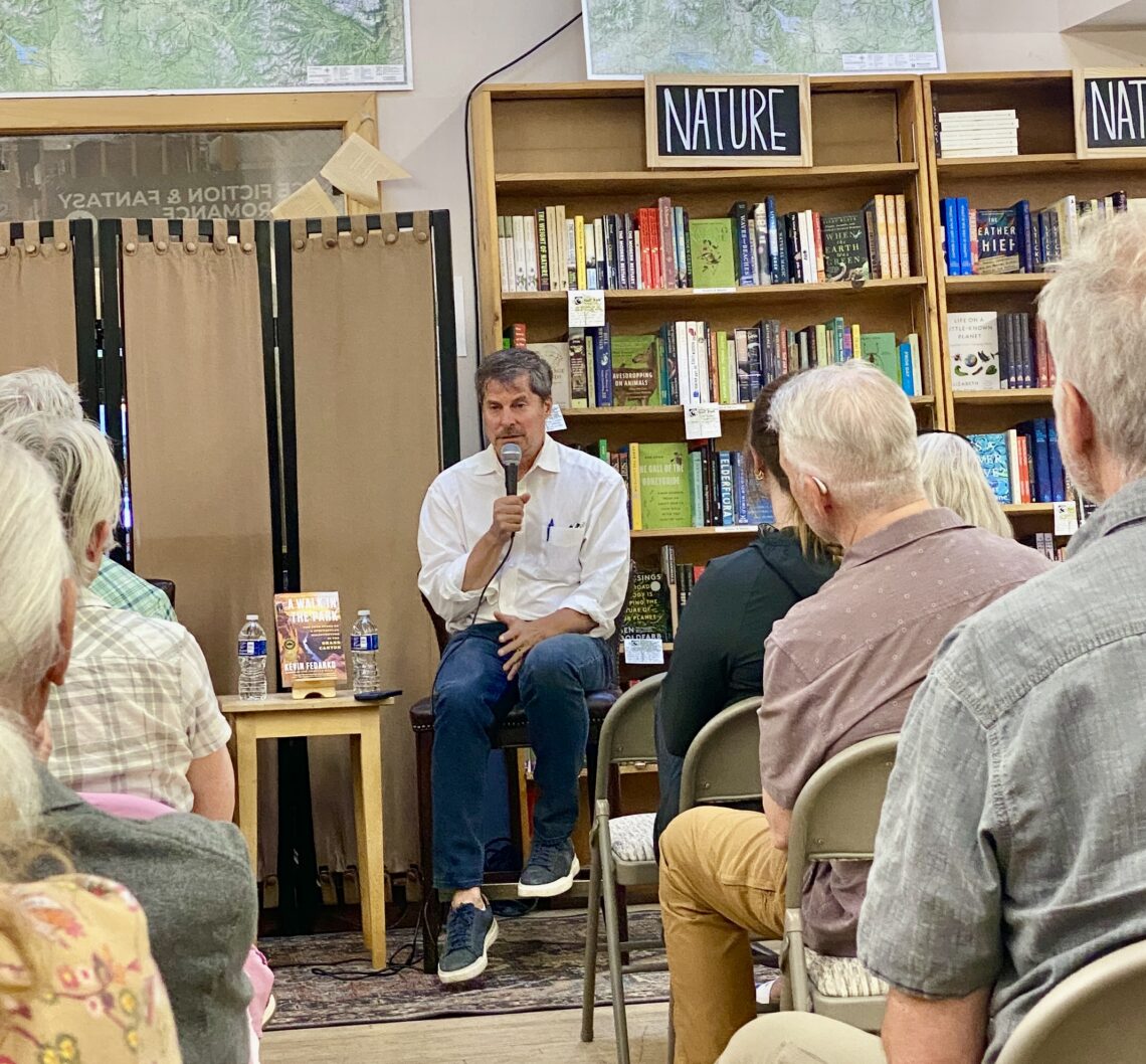 Man in a white shirt holds a microphone while speaking at a seated event in a library or bookstore, shelves of books behind him.