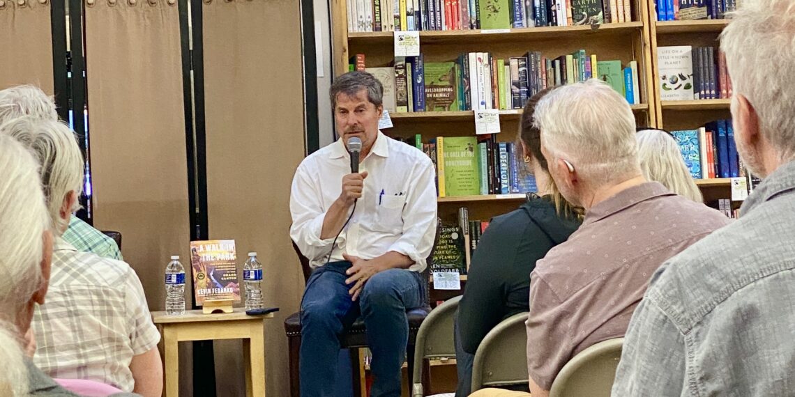 Man in a white shirt holds a microphone while speaking at a seated event in a library or bookstore, shelves of books behind him.