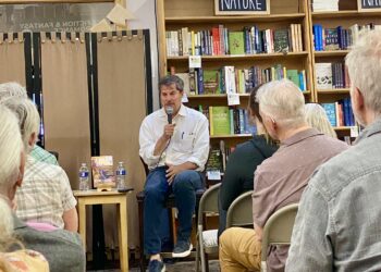 Man in a white shirt holds a microphone while speaking at a seated event in a library or bookstore, shelves of books behind him.