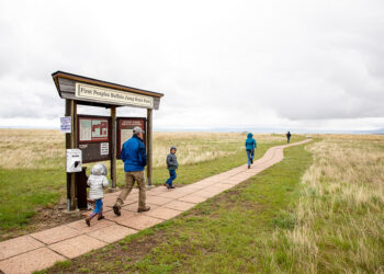 Family walking along a paved trail past a park information kiosk at First Peoples Buffalo Jump State Park, with open prairie and cloudy sky.