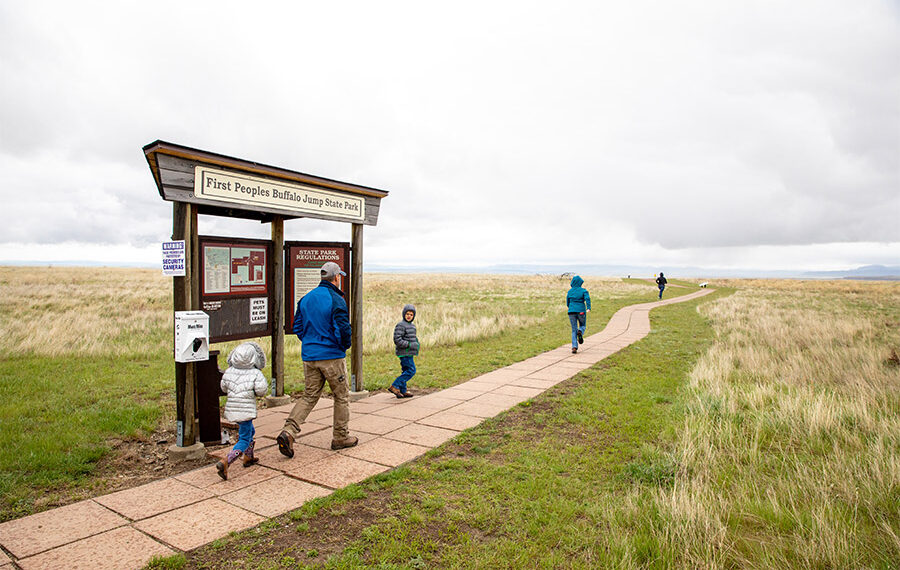 Family walking along a paved trail past a park information kiosk at First Peoples Buffalo Jump State Park, with open prairie and cloudy sky.