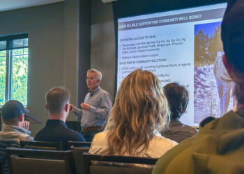 Male presenter speaking to an audience in a conference room with a large slide about community well-being projected on screen.