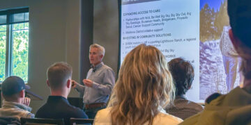 Male presenter speaking to an audience in a conference room with a large slide about community well-being projected on screen.