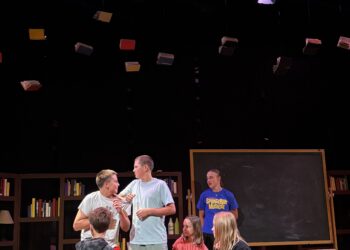 Children on a theater stage with hanging books, shelves, and a chalkboard in a library-like set.