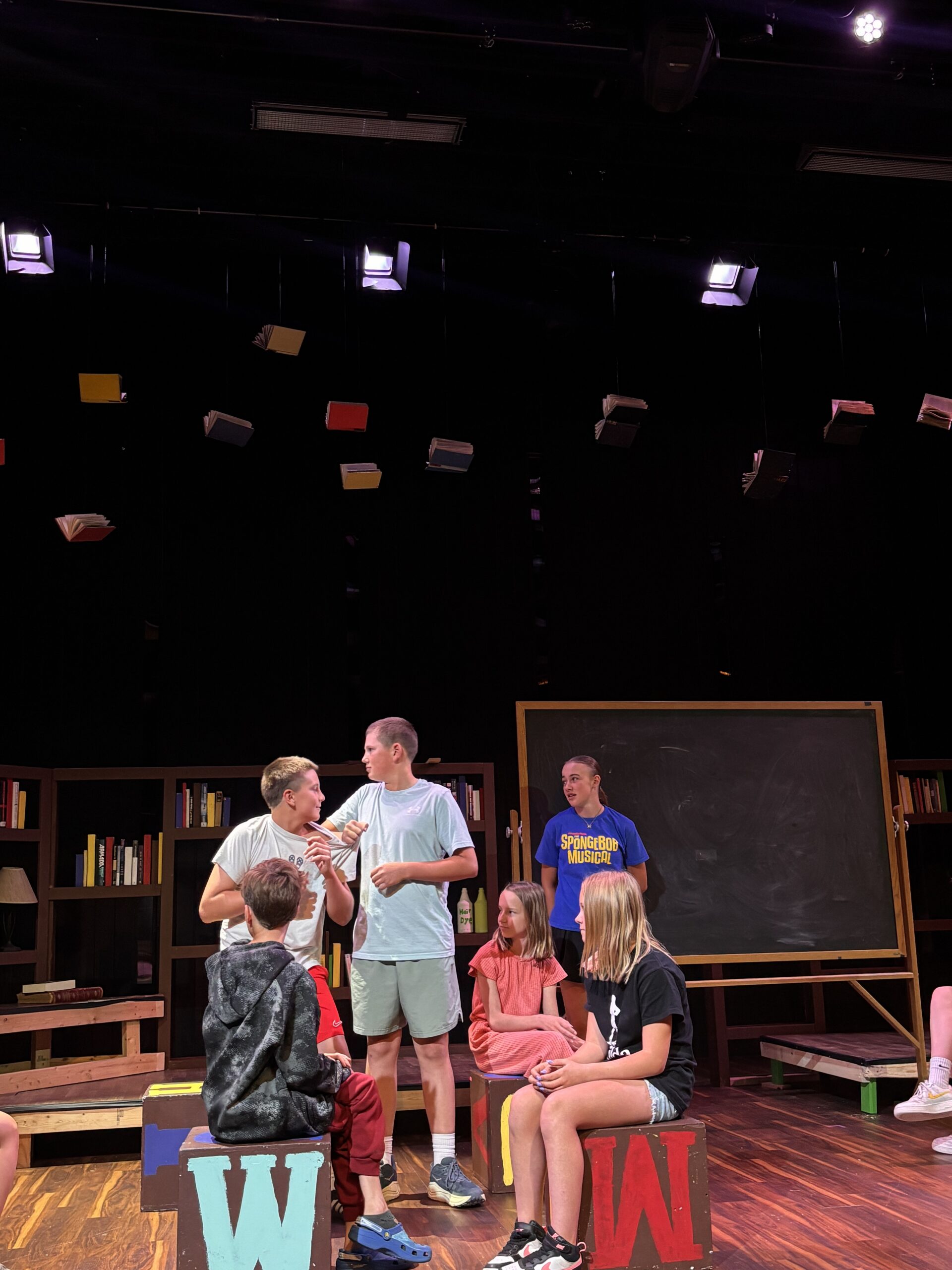 Children on a theater stage with hanging books, shelves, and a chalkboard in a library-like set.