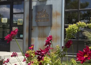 Metal storefront with a rusted corrugated metal wall and a metal sign that reads 'wild crumb' (or similar); bright pink flowers in the foreground.