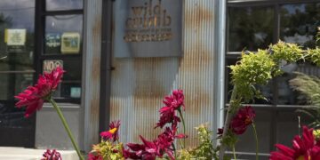 Metal storefront with a rusted corrugated metal wall and a metal sign that reads 'wild crumb' (or similar); bright pink flowers in the foreground.