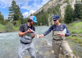 Two people in chest waders stand in a shallow river, exchanging a small bottle while a forested mountain landscape surrounds them.