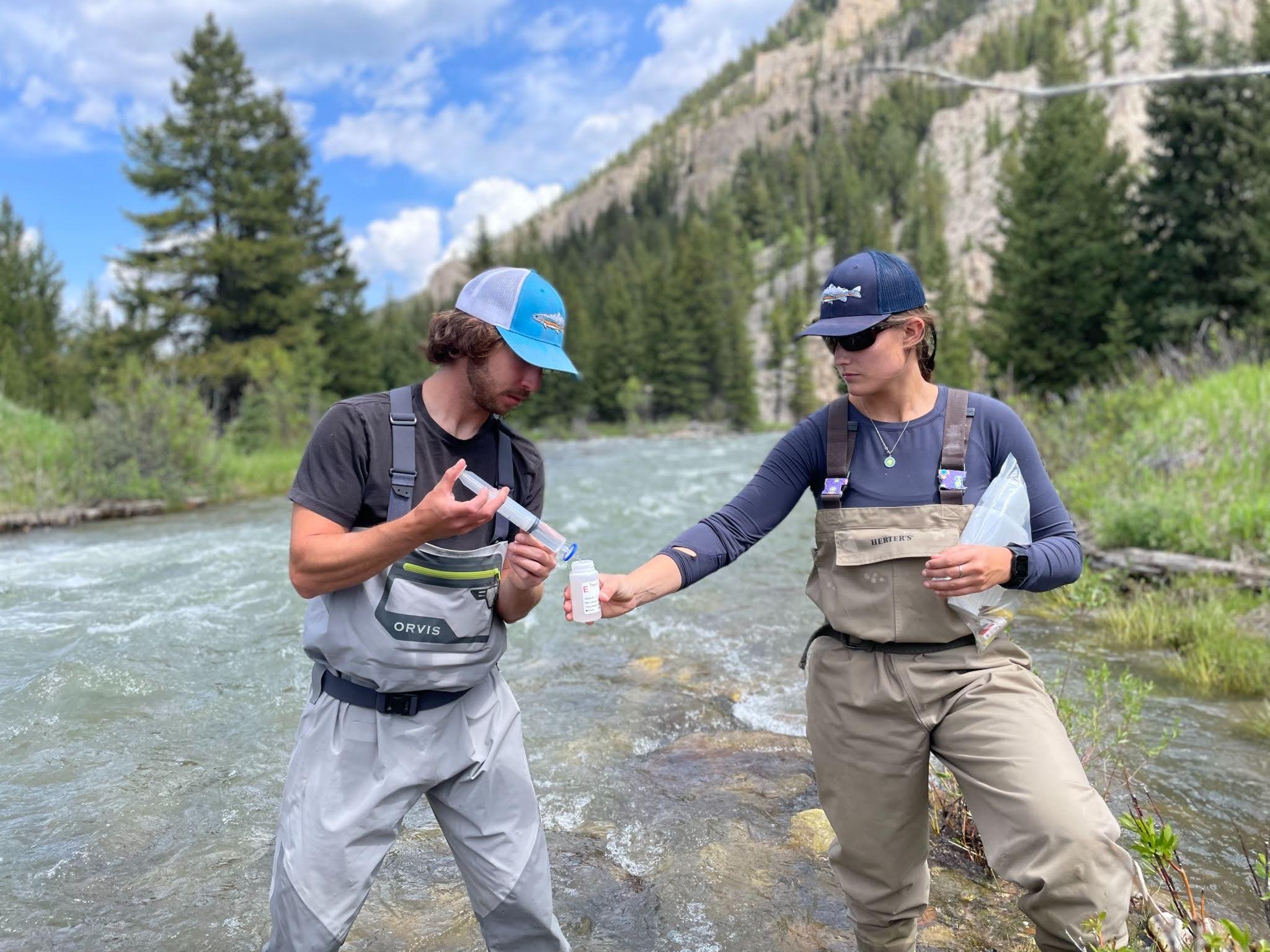 Two people in chest waders stand in a shallow river, exchanging a small bottle while a forested mountain landscape surrounds them.