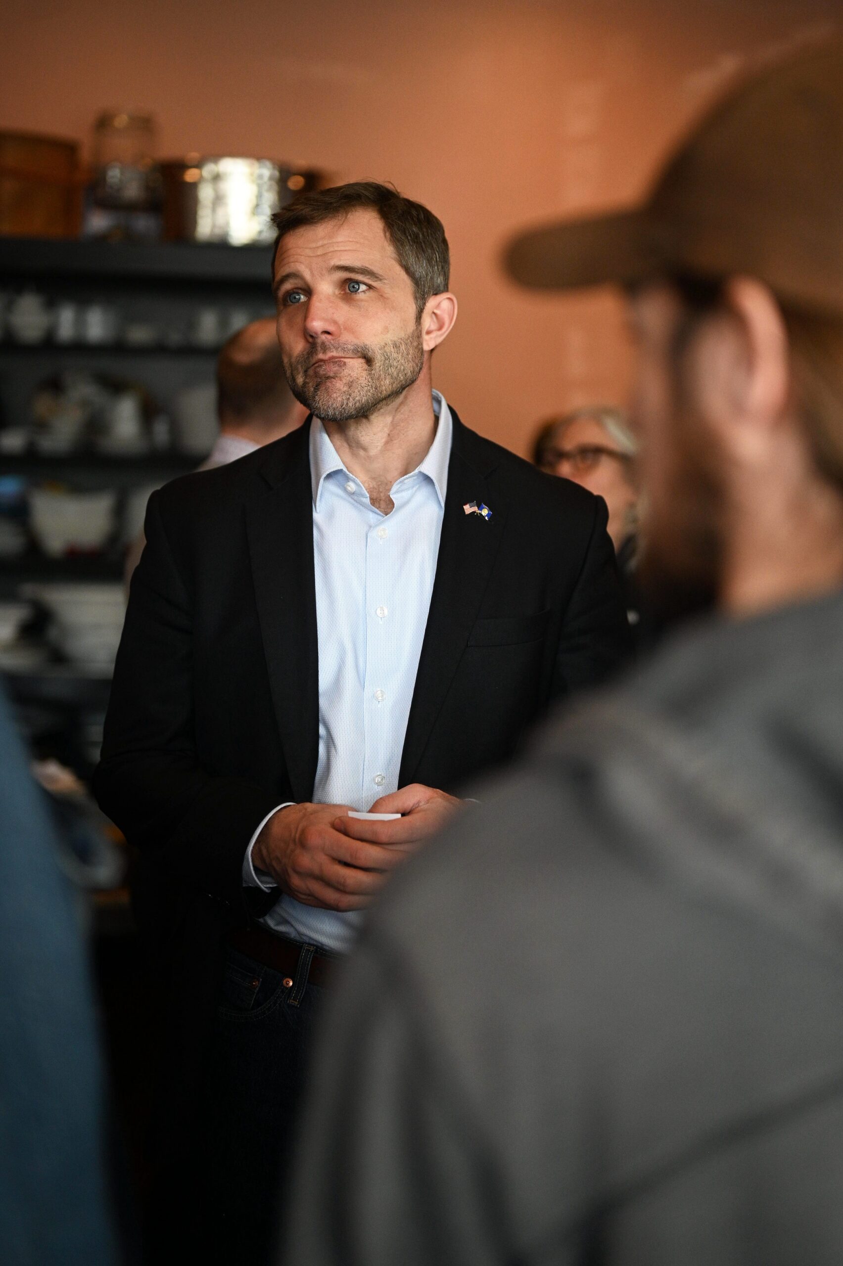 Man in a dark blazer and light blue shirt speaks to a group in a warmly lit room, hands clasped in front of him, attentive expression.