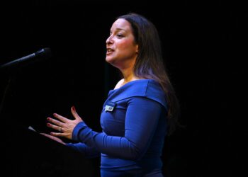 Woman in a blue dress speaks into a microphone on a dark stage, with a name badge visible on her chest.