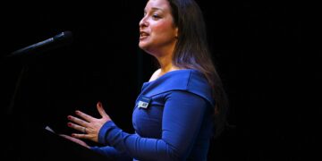 Woman in a blue dress speaks into a microphone on a dark stage, with a name badge visible on her chest.