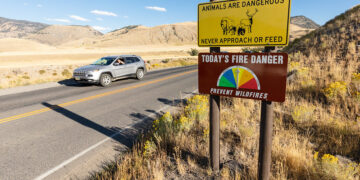 Desert highway with wildlife and fire danger signs; a gray SUV drives past on a two-lane road with dry vegetation nearby.