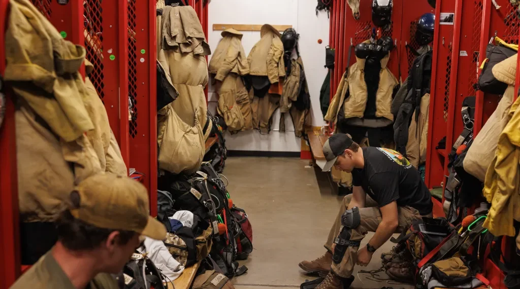 Trainee firefighter sits on a bench in a red locker room, tying turnout boots amid hanging coats, helmets, and gear.