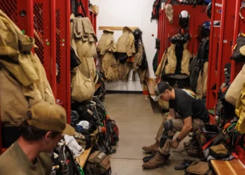 Trainee firefighter sits on a bench in a red locker room, tying turnout boots amid hanging coats, helmets, and gear.