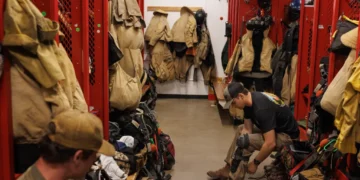 Trainee firefighter sits on a bench in a red locker room, tying turnout boots amid hanging coats, helmets, and gear.