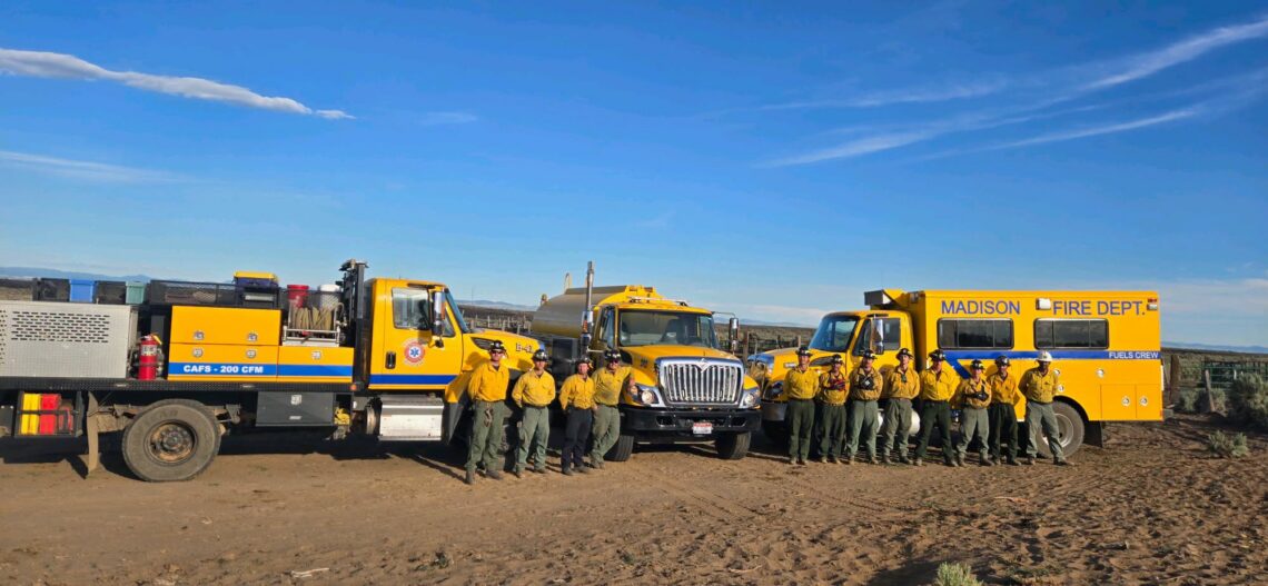 A line of yellow fire trucks with firefighters in uniform standing in front of them under a blue sky in a desert setting. Madison Fire Dept visible on the large yellow vehicle on the right.