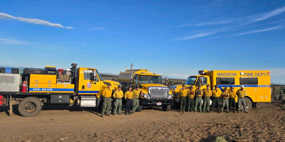 A line of yellow fire trucks with firefighters in uniform standing in front of them under a blue sky in a desert setting. Madison Fire Dept visible on the large yellow vehicle on the right.