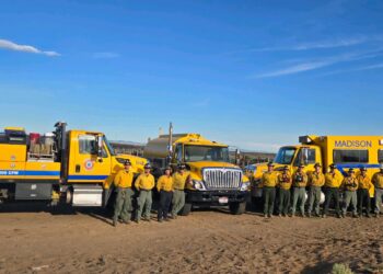 A line of yellow fire trucks with firefighters in uniform standing in front of them under a blue sky in a desert setting. Madison Fire Dept visible on the large yellow vehicle on the right.