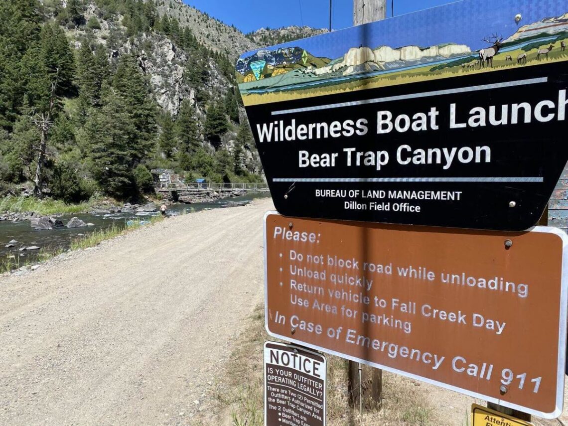 Sign for Wilderness Boat Launch at Bear Trap Canyon beside a dirt road, river and pine-covered canyon backdrop.