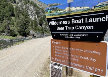Sign for Wilderness Boat Launch at Bear Trap Canyon beside a dirt road, river and pine-covered canyon backdrop.