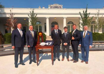 Six men in business suits pose outside a white columned building with an award display and American flags behind them.