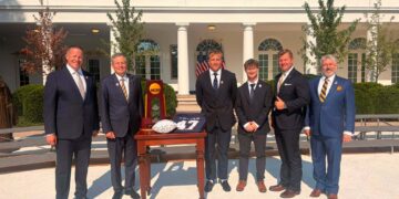 Six men in business suits pose outside a white columned building with an award display and American flags behind them.