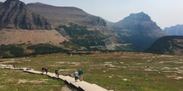 Hikers walk along a wooden boardwalk through a grassy alpine valley with rocky mountains in the distance under a clear sky.