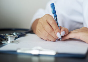 Person wearing white coat writes on paper with a blue pen; stethoscope lies on the desk nearby, suggesting a medical setting.