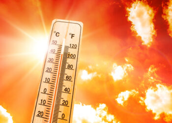 Thermometer in the foreground showing high temperature against a blazing orange sky with sun glare.
