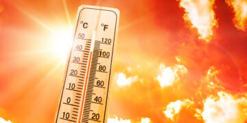 Thermometer in the foreground showing high temperature against a blazing orange sky with sun glare.