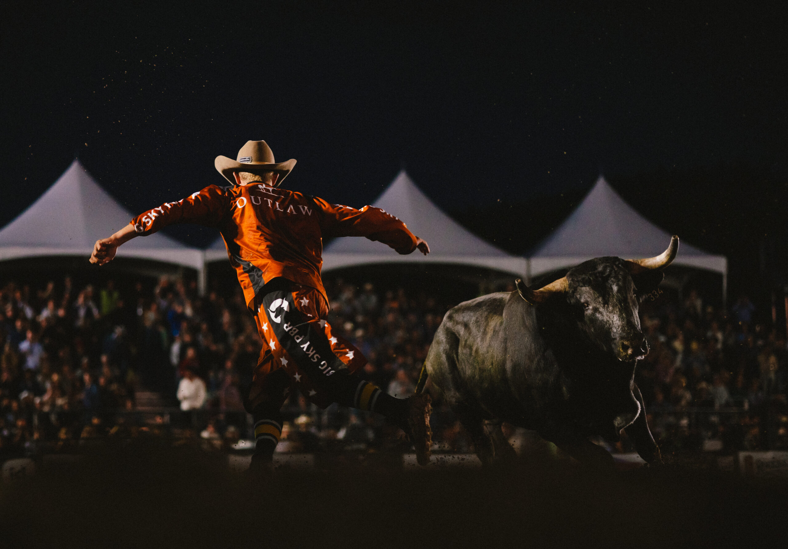 Rodeo rider in an orange suit and cowboy hat jumps off a bucking bull at night in front of a cheering crowd.