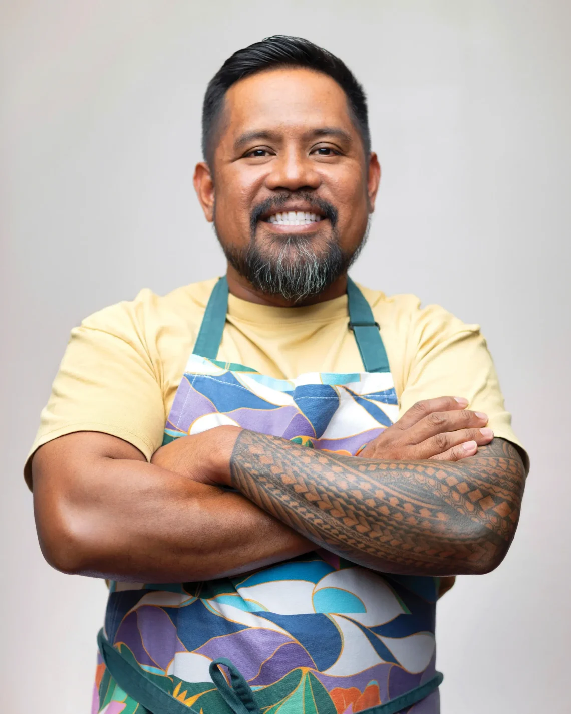 Smiling man with a trimmed beard and tattooed forearm wearing a colorful apron, arms crossed in a portrait against a neutral background.