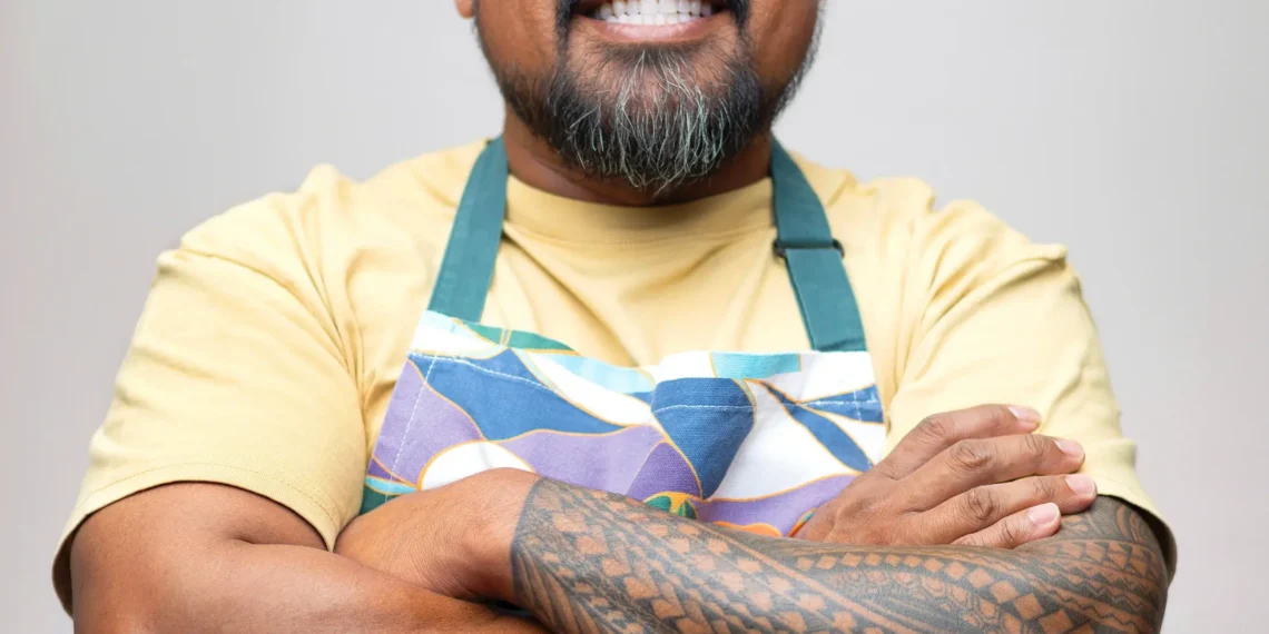Smiling man with a trimmed beard and tattooed forearm wearing a colorful apron, arms crossed in a portrait against a neutral background.