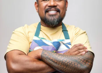 Smiling man with a trimmed beard and tattooed forearm wearing a colorful apron, arms crossed in a portrait against a neutral background.